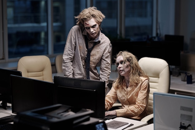 Premium Photo | Young spooky zombie businessman standing next to female colleague sitting by desk in front of computer monitor during teamwork